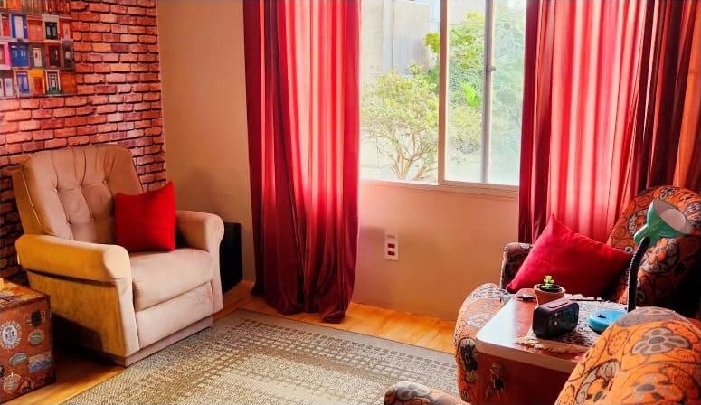 Poltrona bege junto à parede de tijolos, ao lado da janela com cortinas vermelhas e tapete no chão de madeira.