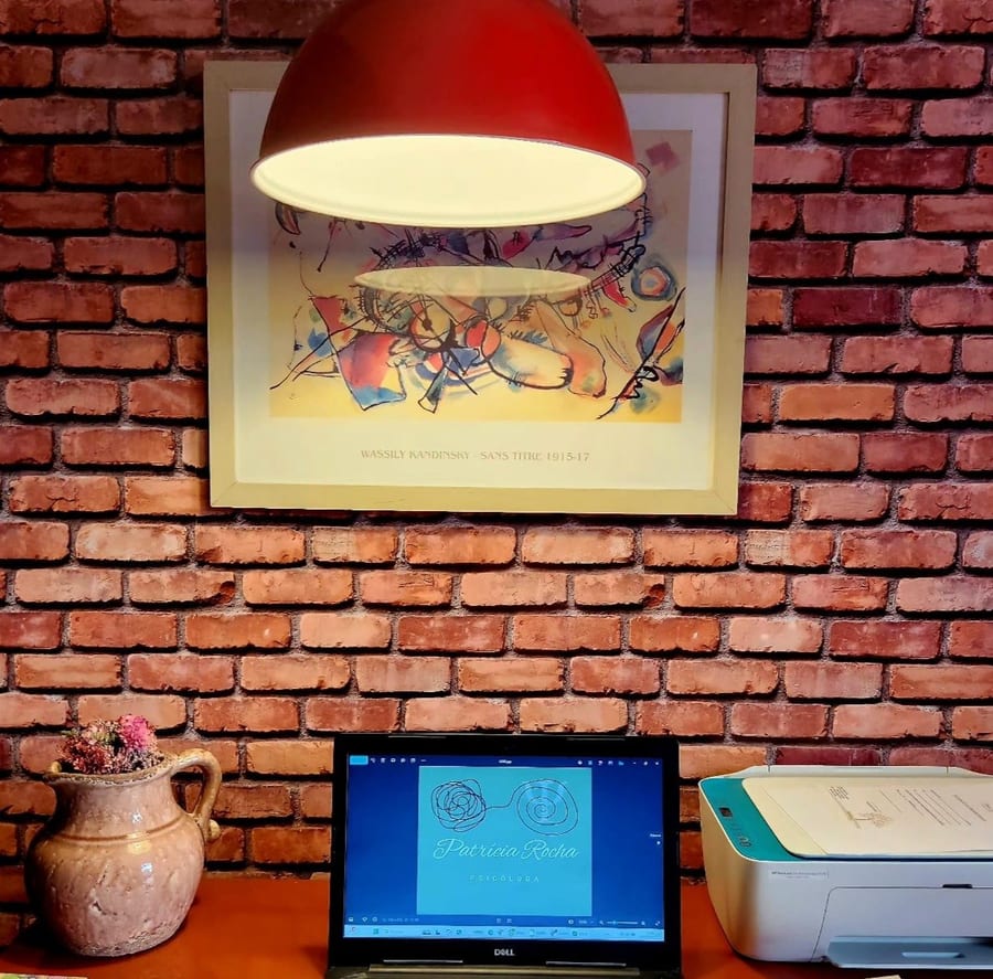 Mesa de trabalho com computador, cadernos e uma jarra de cerâmica, sob a luminária vermelha e uma gravura de Kandinsky na parede de tijolos.