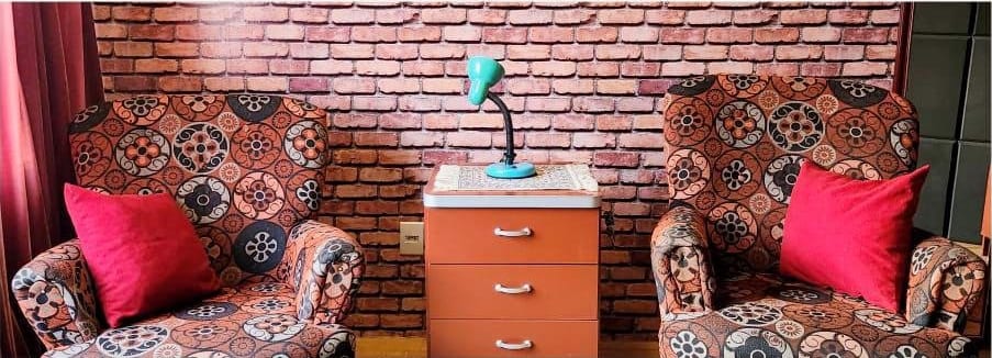 Duas poltronas estampadas, uma de frente para a outra, com almofadas vermelhas e uma cômoda entre elas diante da parede de tijolos.