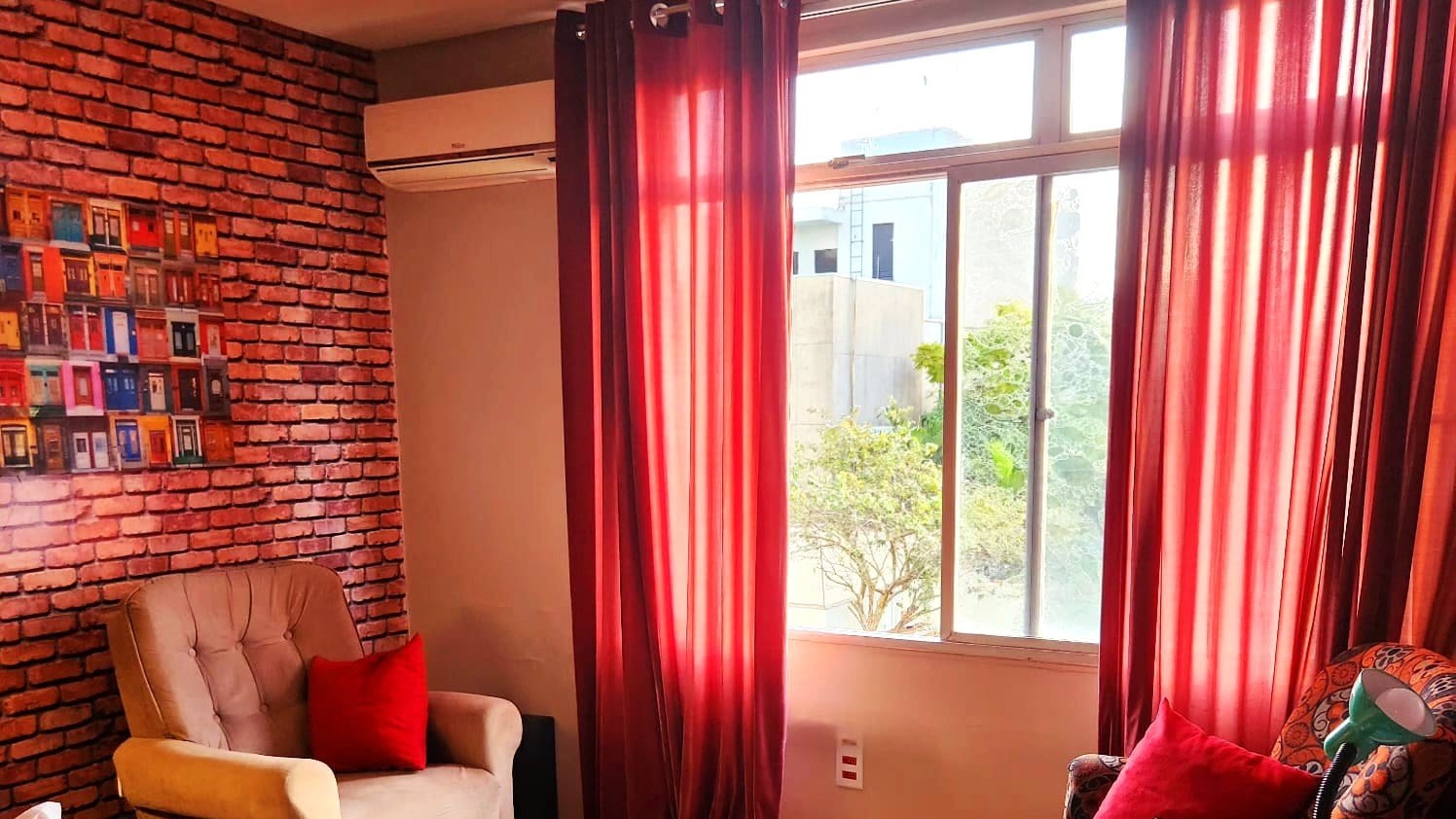 Sala de atendimento com cortinas vermelhas abertas, janela com vista para uma árvore e poltronas em torno de uma mesa baixa.